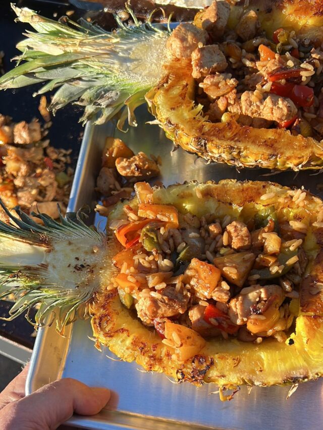 Stuffed pineapple halves with fried rice, vegetables, and diced chicken, served on a metal tray.