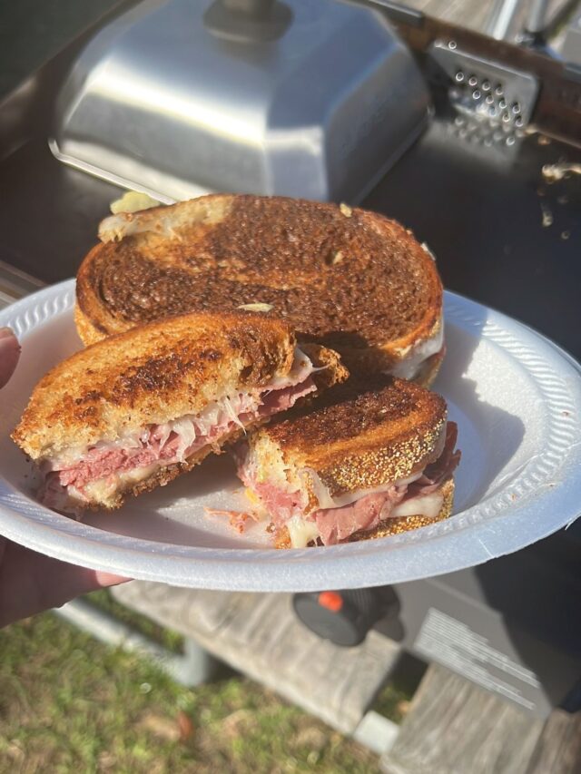 A person holds a plate with a grilled Reuben sandwich cut in half, showcasing melted cheese and layers of ham inside, with a Blackstone grill in the background.