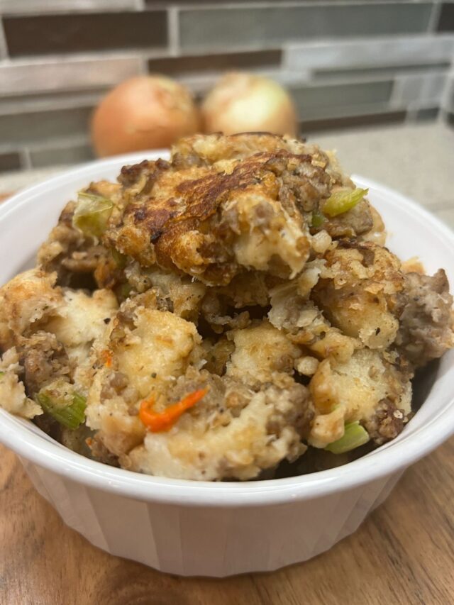 A bowl of traditional sausage stuffing with visible chunks of celery, carrot, and onion, set against a kitchen backsplash.