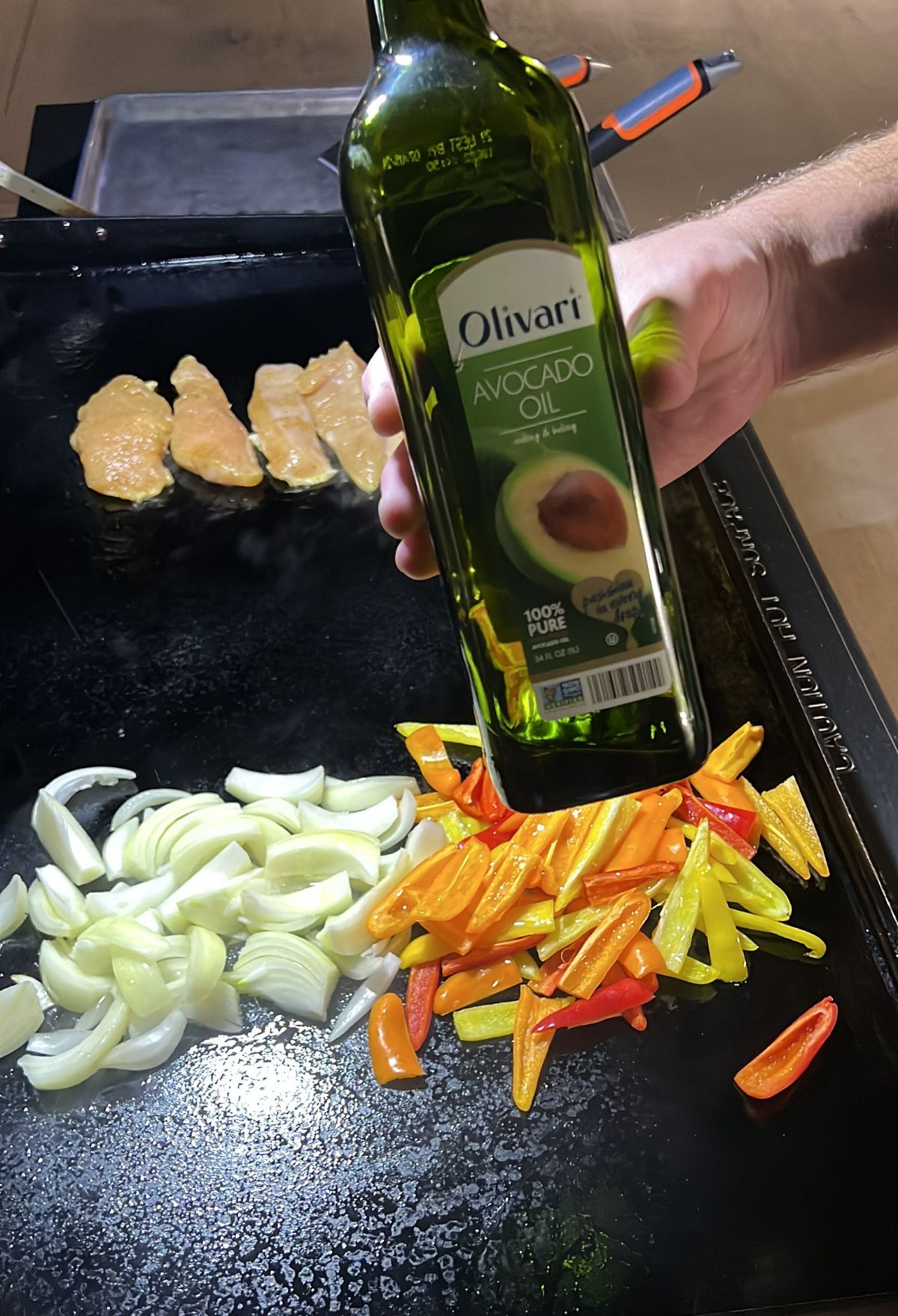 A person pouring avocado oil onto a griddle with sliced onions and bell peppers, with chicken pieces cooking in the background.