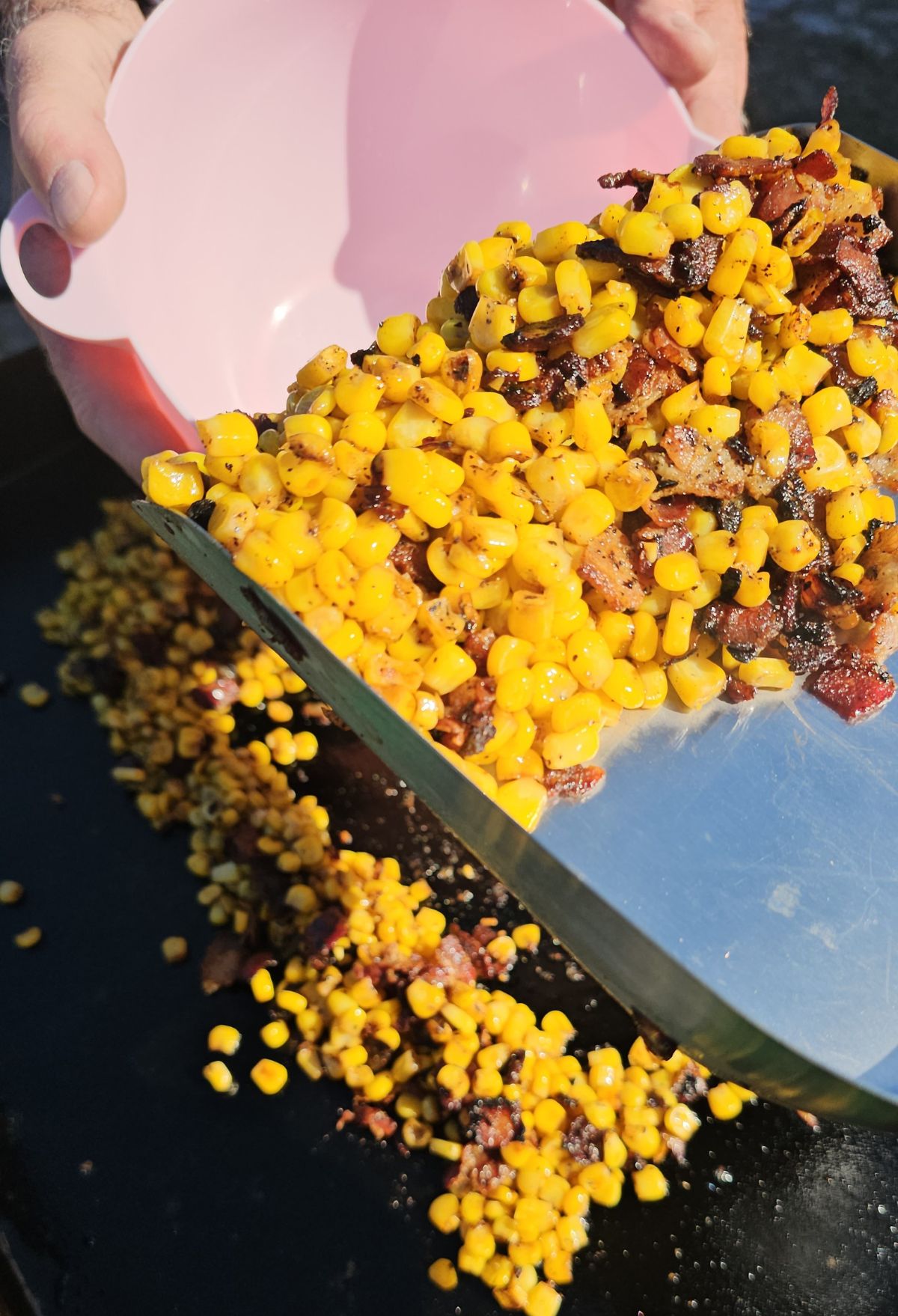 A person accidentally spills a mixture of corn and bacon from a pink bowl onto a cooking surface.