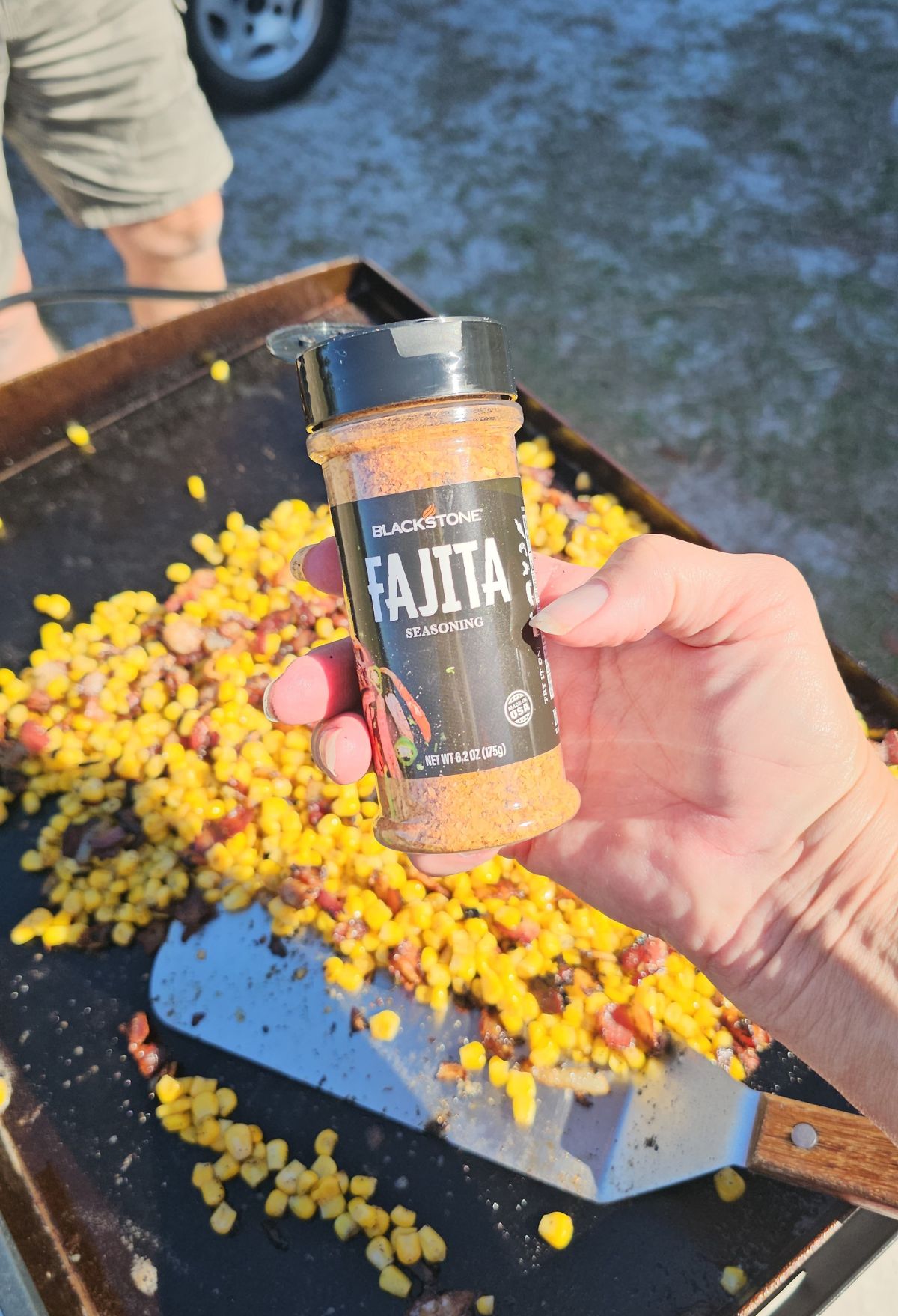 A person holding a container of fajita seasoning over a griddle full of cooking corn and peppers.