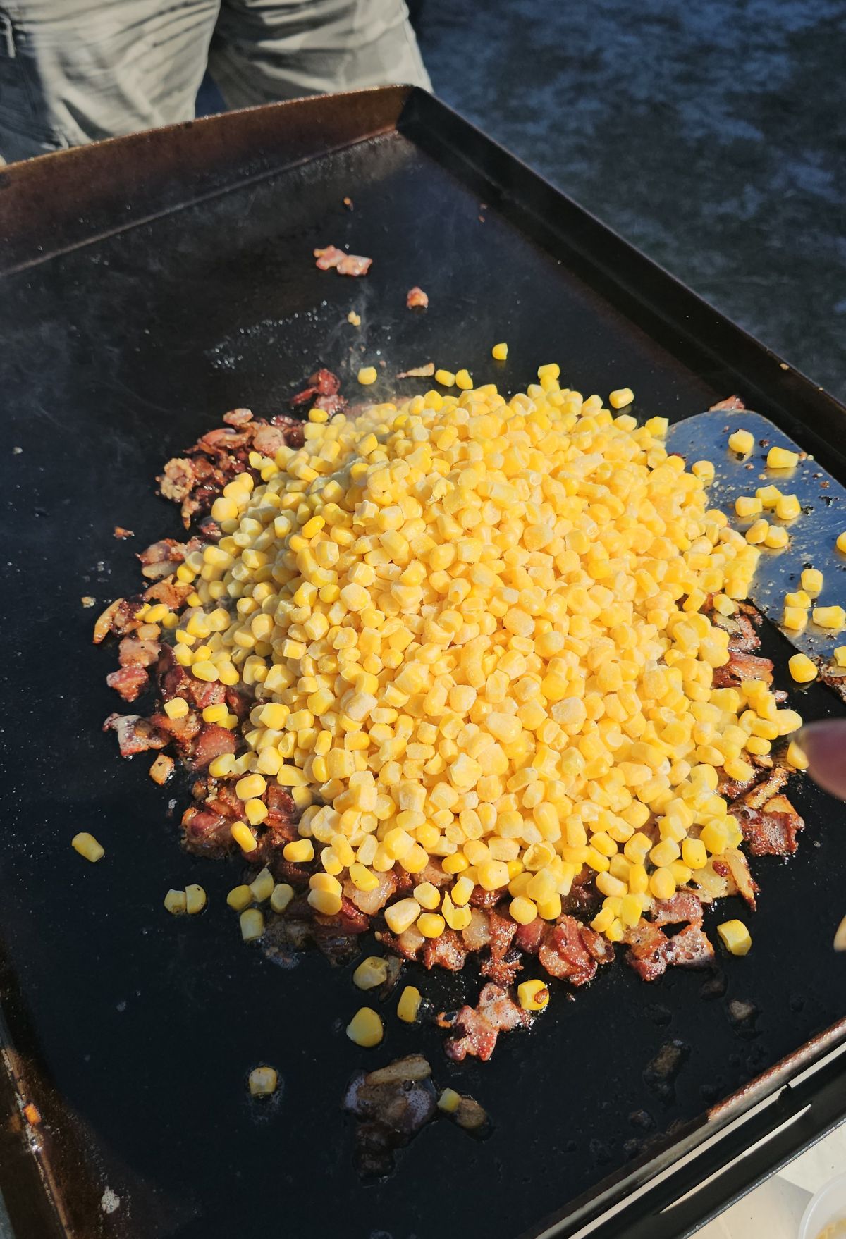 Corn kernels cooking with bacon pieces on a flat grill.