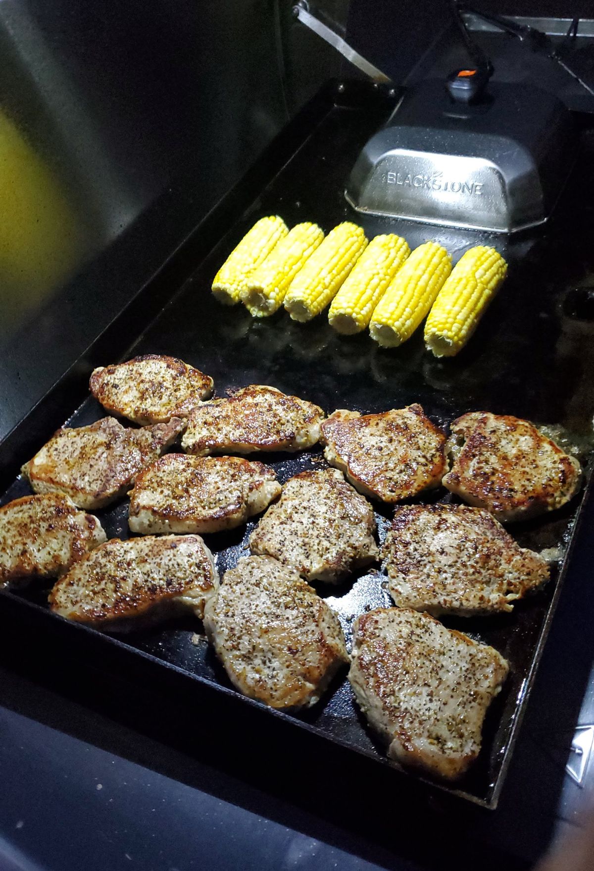 Seasoned pork chops and corn on the cob grilling on a flat top grill.