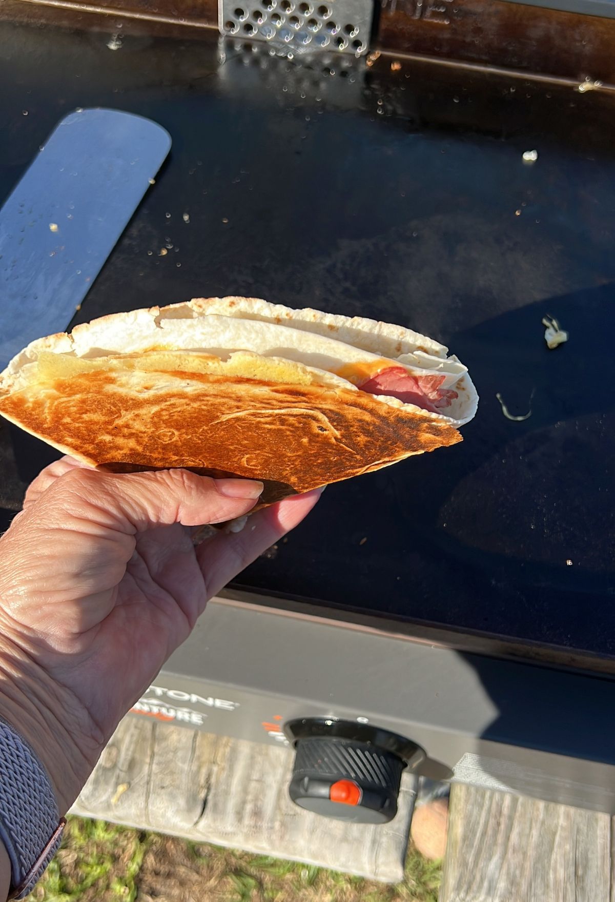 Hand holding an overstuffed sandwich with visible eggs and meats on a griddle background.