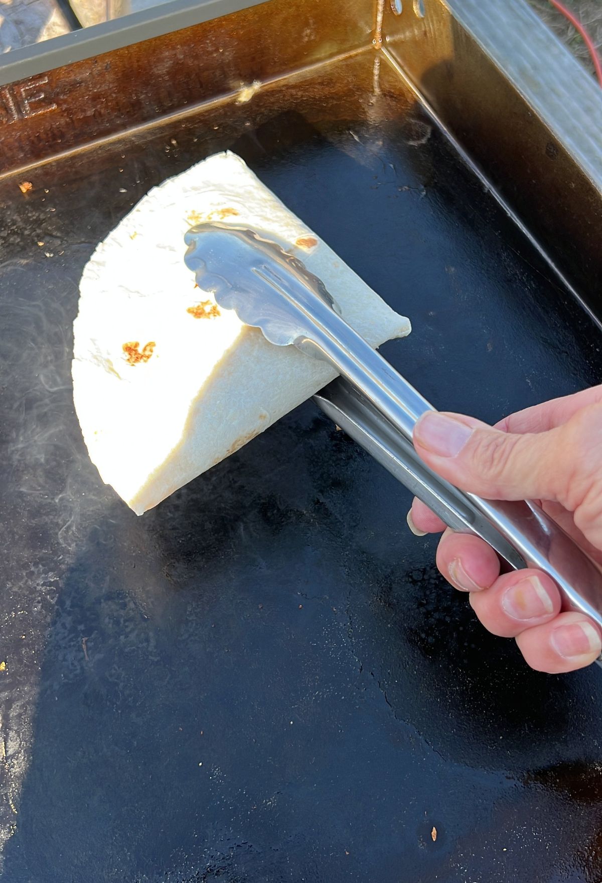 Grilling a tortilla on an outdoor flat-top grill using metal tongs.