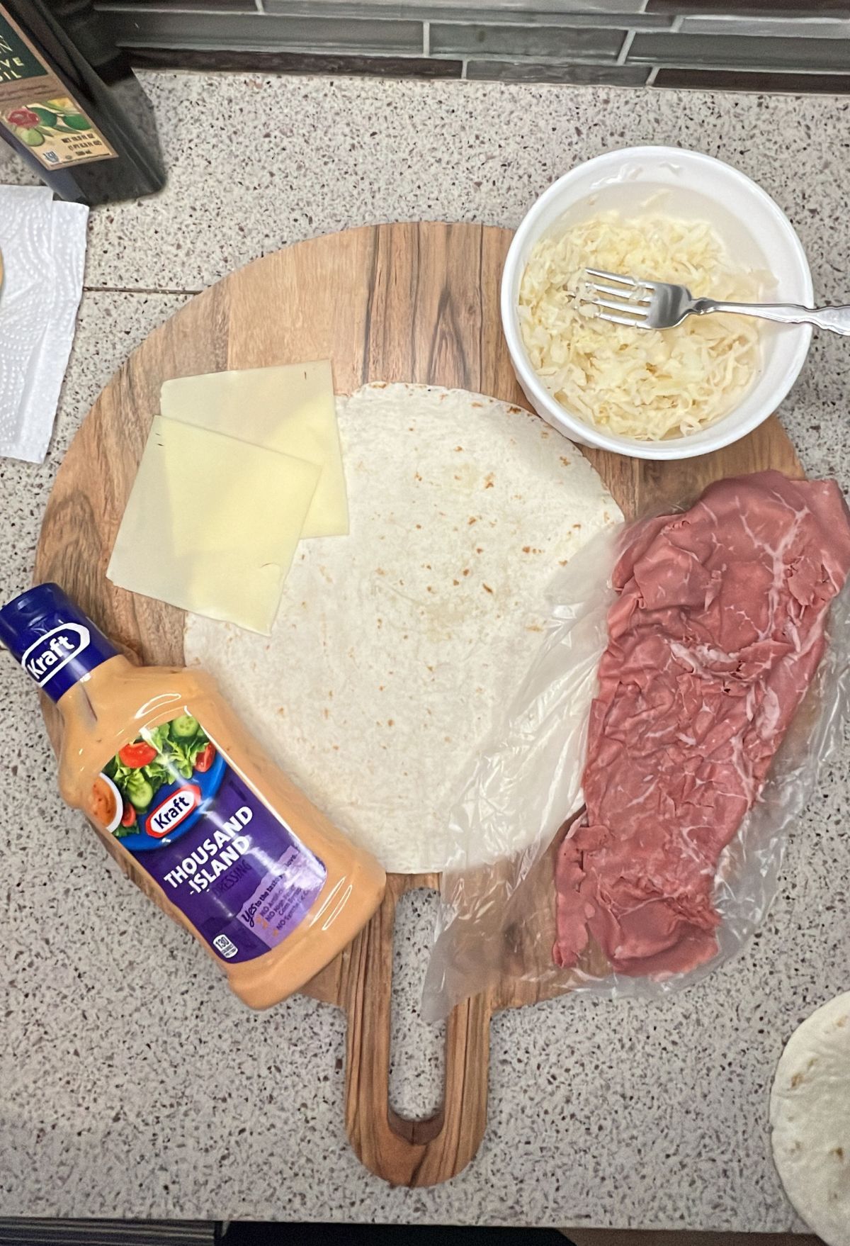 Ingredients for a meal laid out on a cutting board, including sliced beef, cheese slices, tortillas, mayonnaise, and noodles.