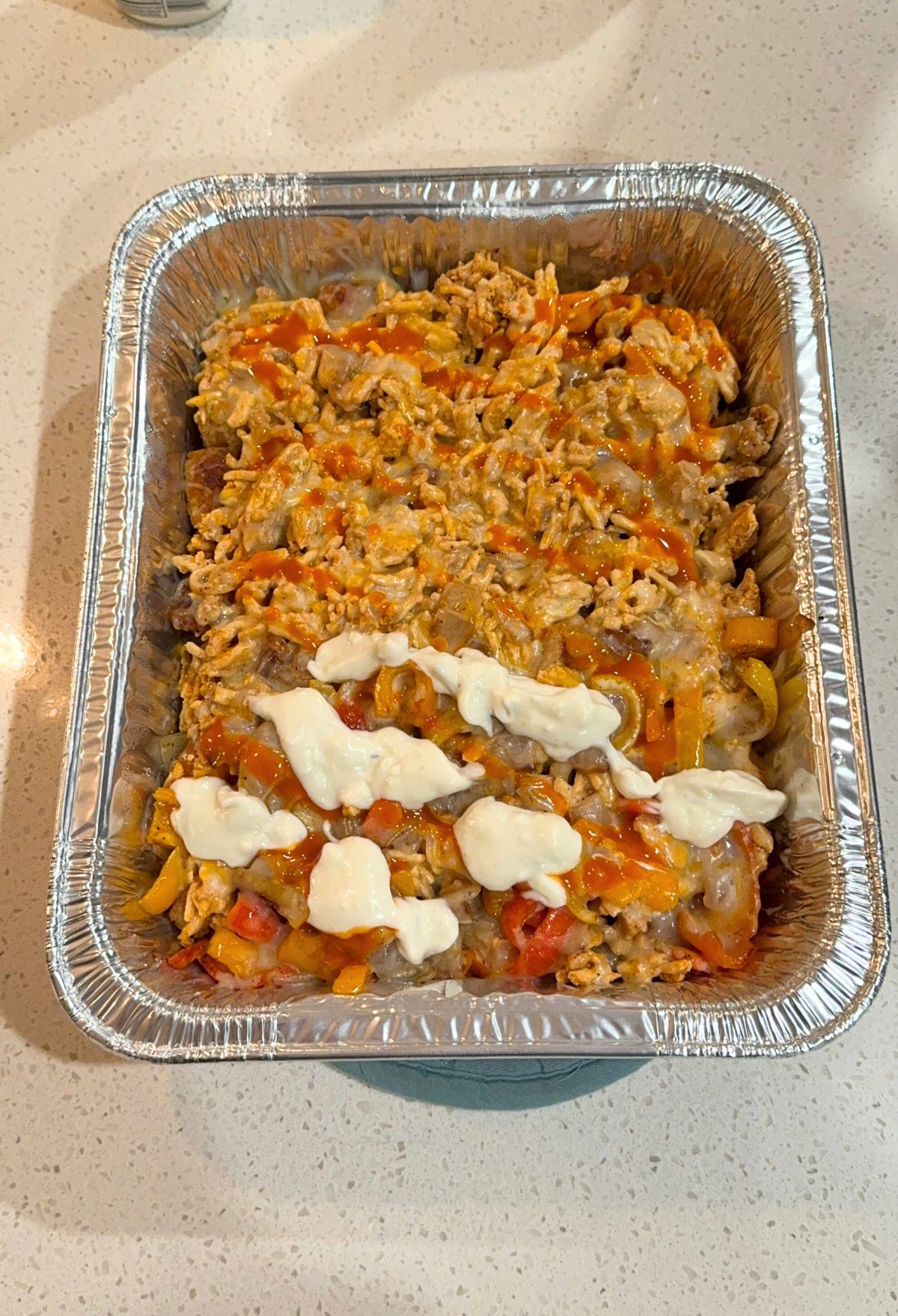 Aluminum tray containing a baked pasta dish with tomato chunks, melted cheese, and dollops of white sauce on top.