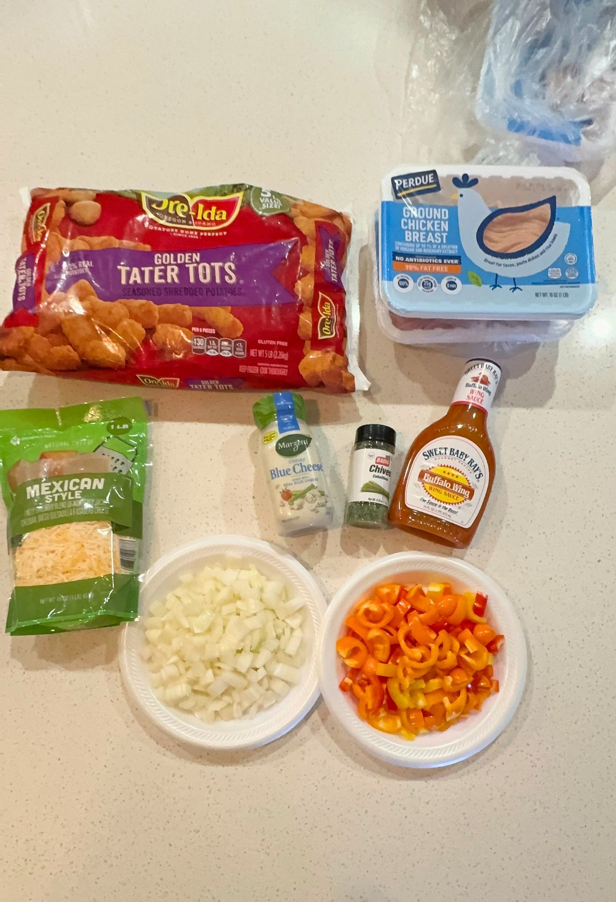 Ingredients for a meal preparation displayed on a kitchen counter, including tater tots, chicken breast, cheese, vegetables, and condiments.