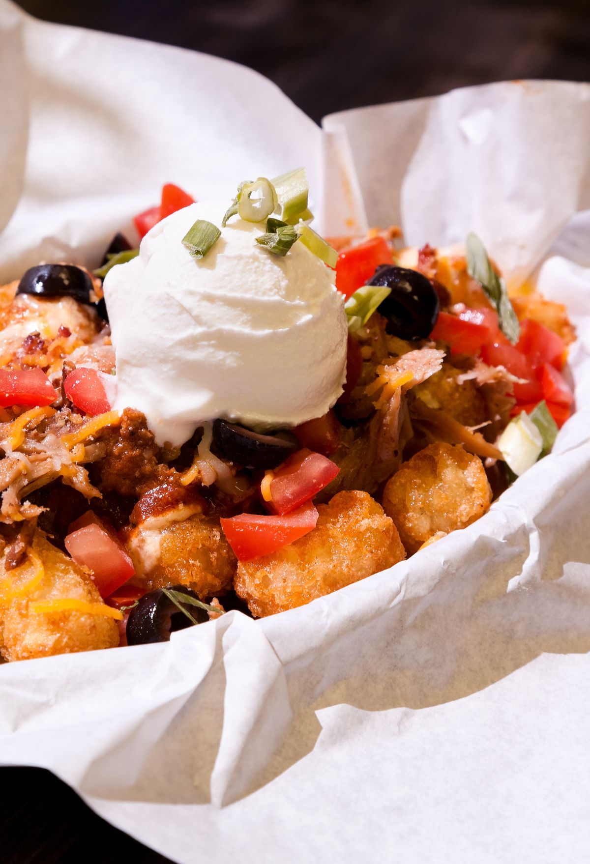 A serving of loaded tater tots topped with ground beef, diced tomatoes, black olives, green onions, and a dollop of sour cream.