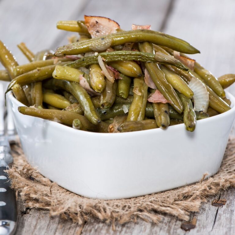 A bowl of cooked green beans with onions and bacon bits on a rustic wooden surface.