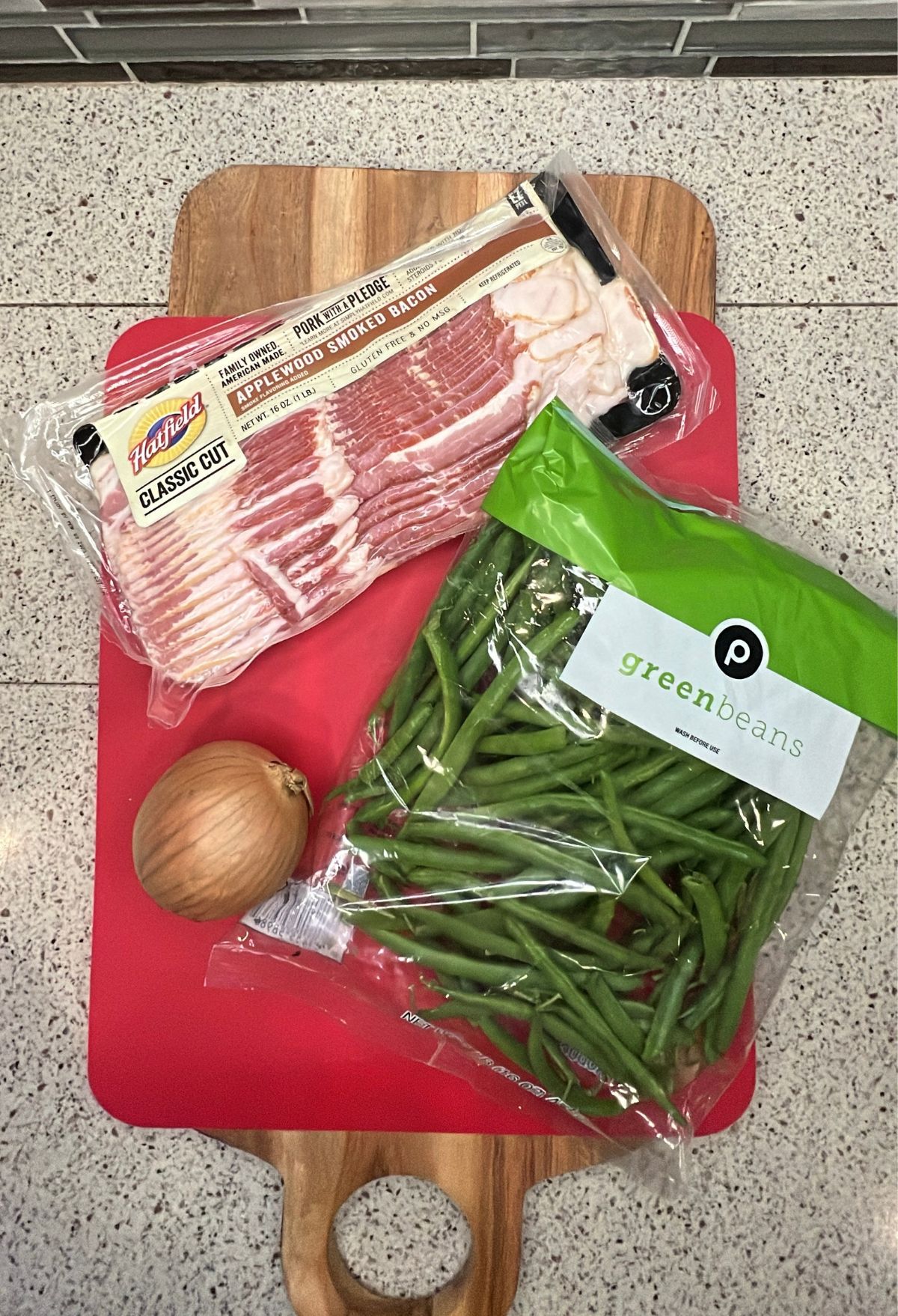 Bacon, an onion, and a bag of green beans on a red cutting board.