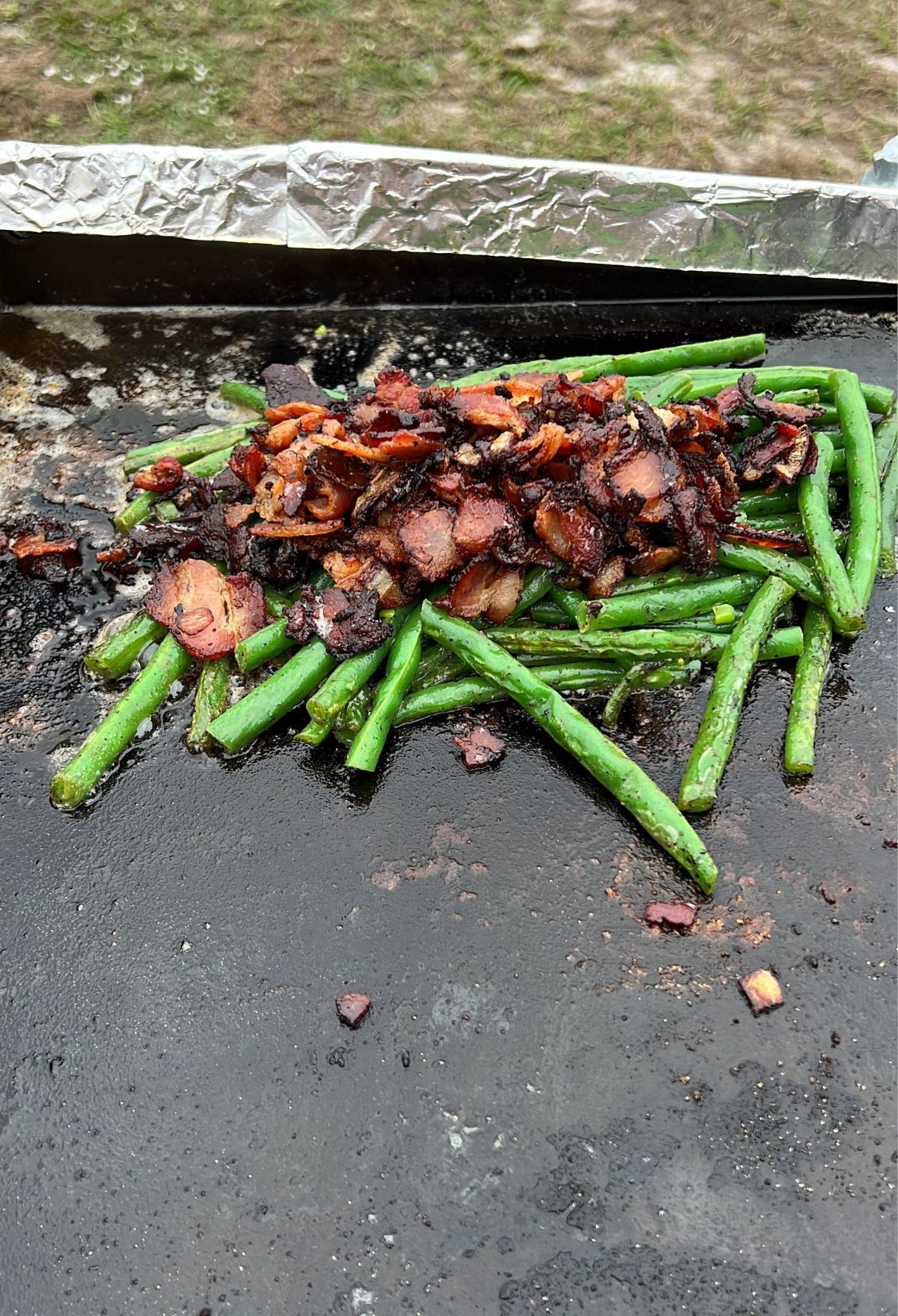 Grilled green beans and chopped bacon cooking on an outdoor flat grill, with visible seasoning.