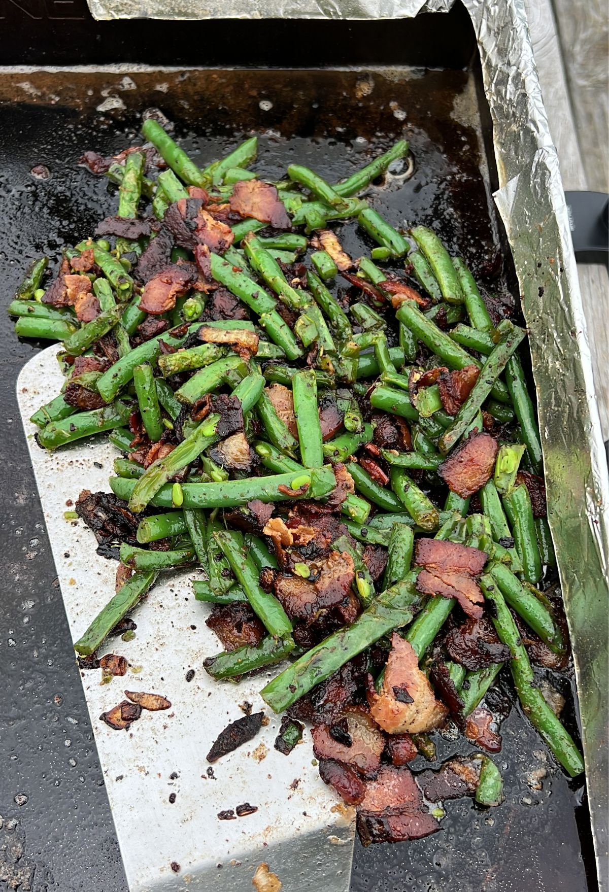 Cooked green beans and bacon pieces on a griddle.