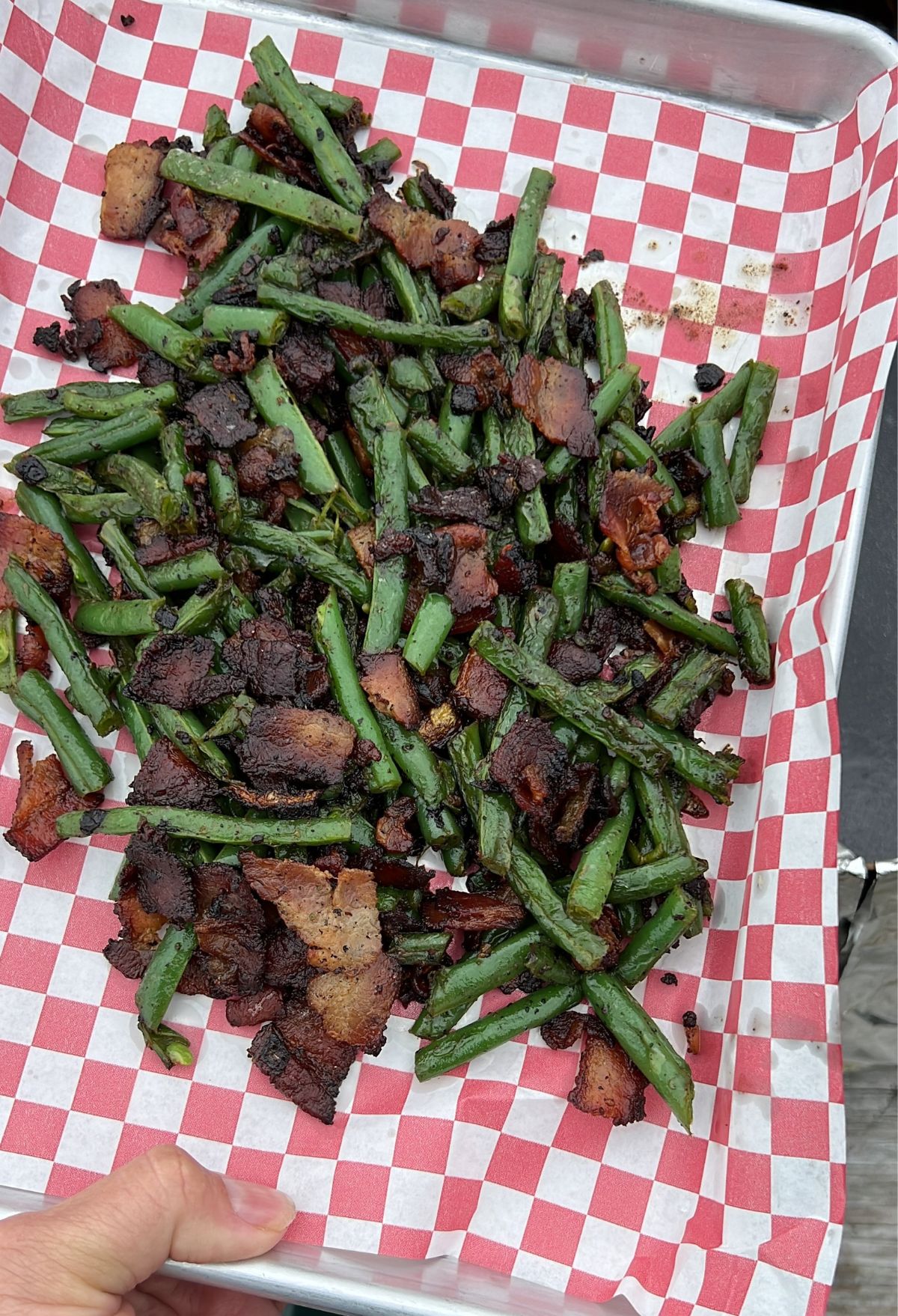 A tray of sautéed green beans and bacon served on red and white checkered paper, held by a person.