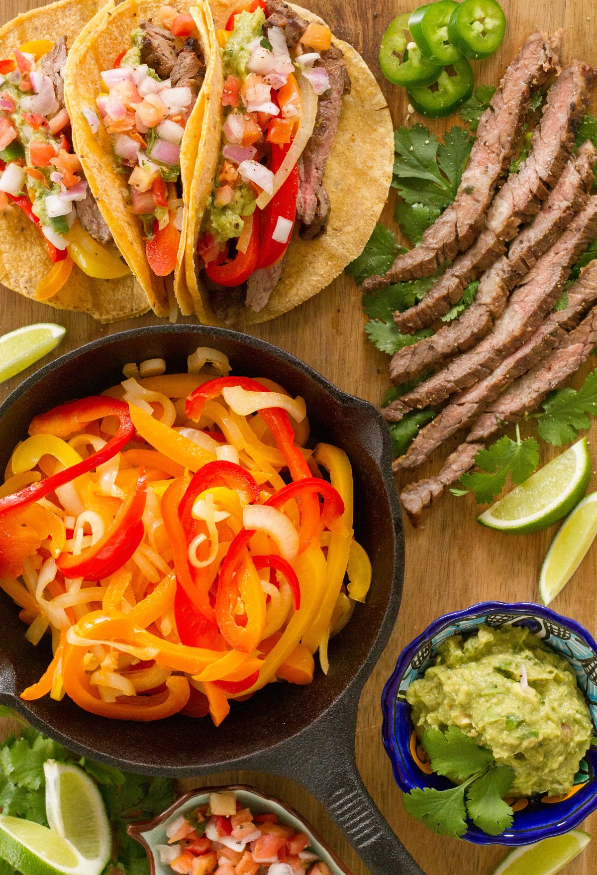 A variety of mexican dishes including tacos, sliced steak, a skillet of sautéed bell peppers, guacamole, and fresh limes on a wooden table.