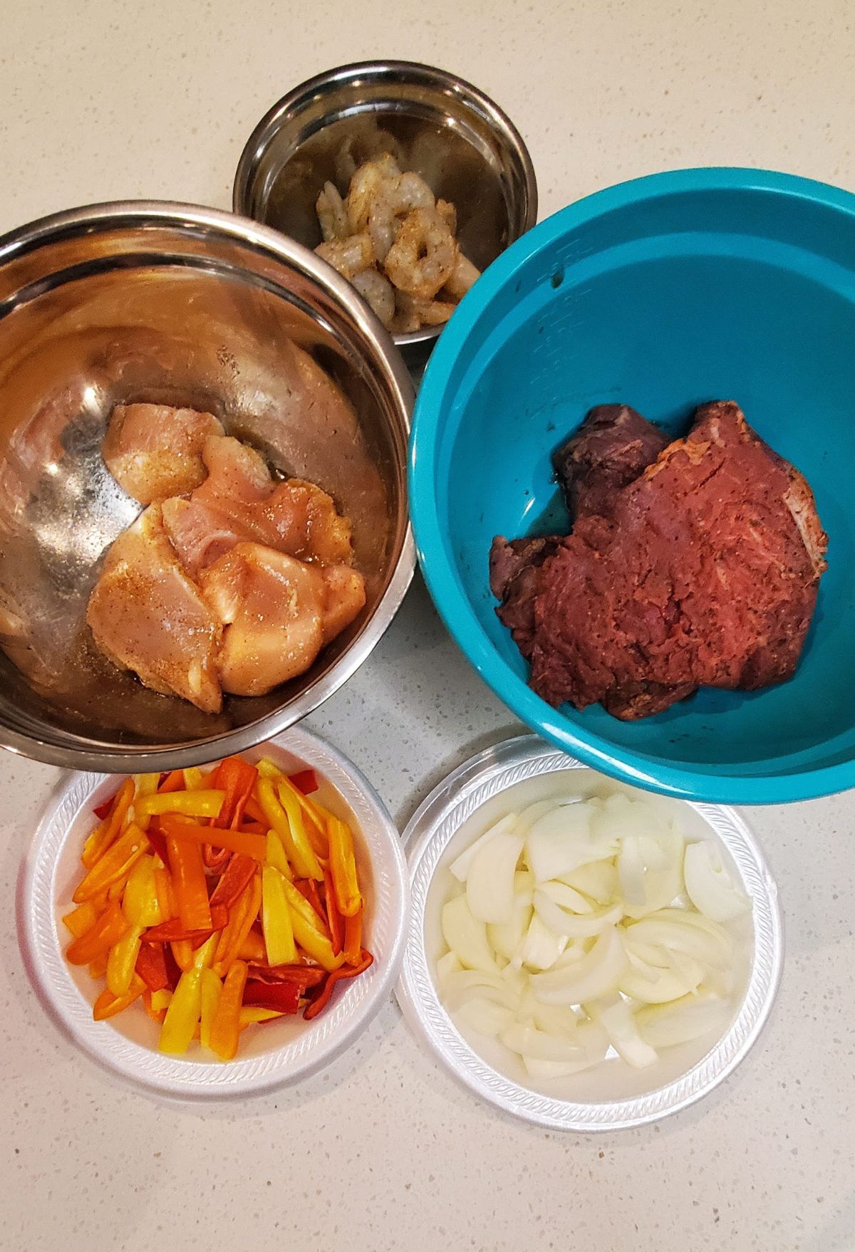 Ingredients for cooking laid out on a counter: seasoned chicken, shrimp, ground beef, sliced bell peppers, and onions in separate bowls.