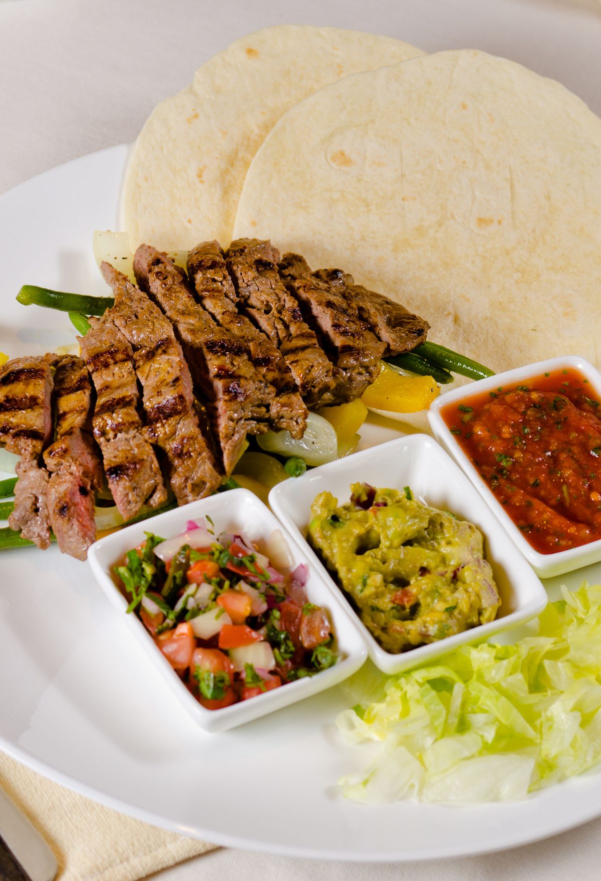 Grilled steak fajitas with tortillas, guacamole, salsa, and vegetables on a white plate.