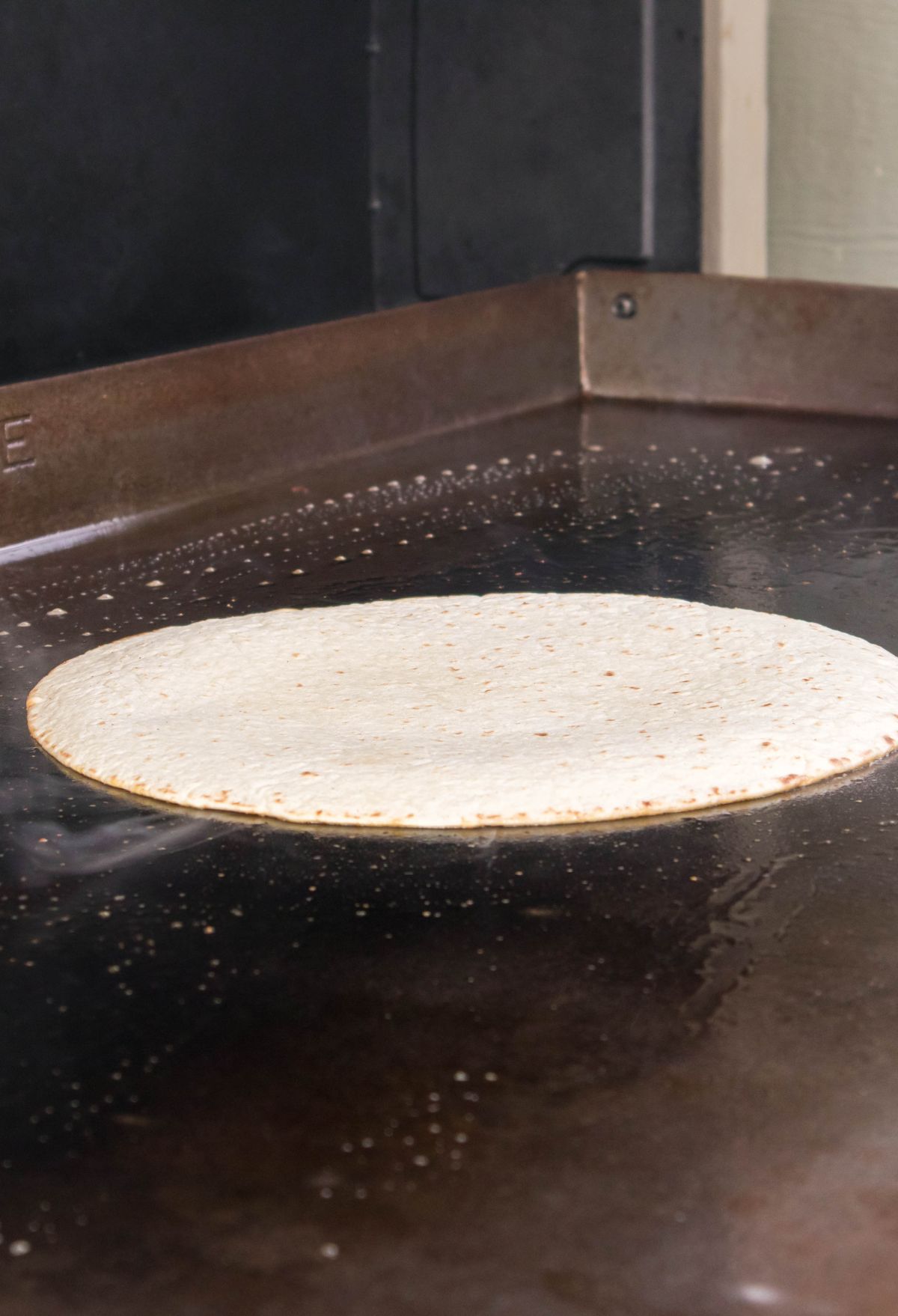 A tortilla cooking on a large, heated flat griddle, with visible steam rising from its surface.