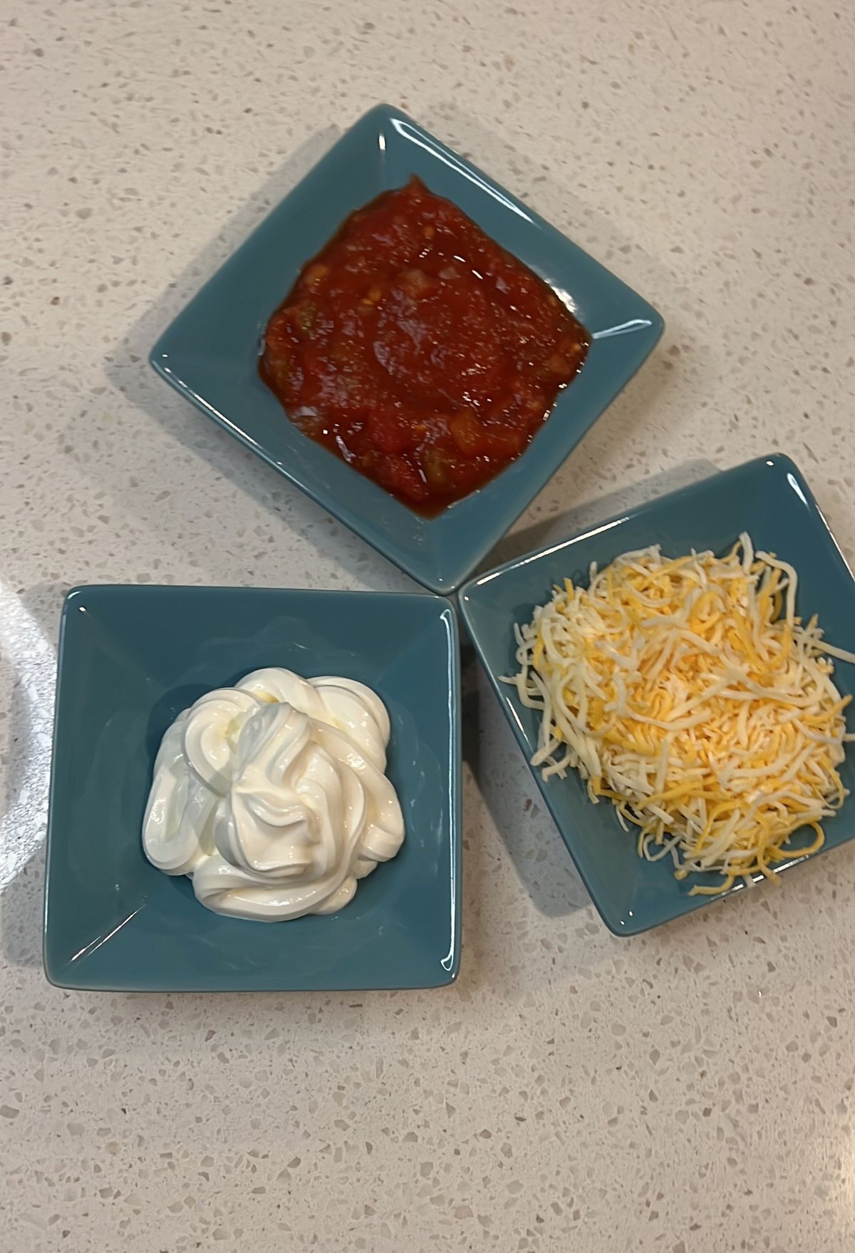 Three square bowls on a countertop containing salsa, sour cream, and shredded cheese respectively.