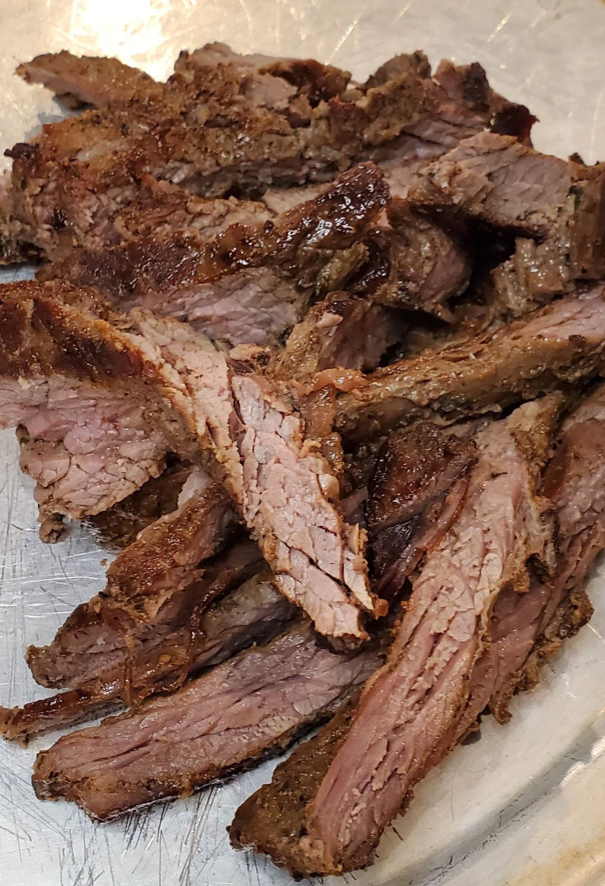 Sliced grilled steak on a metal tray, showing juicy, medium-cooked meat with a light brown crust.