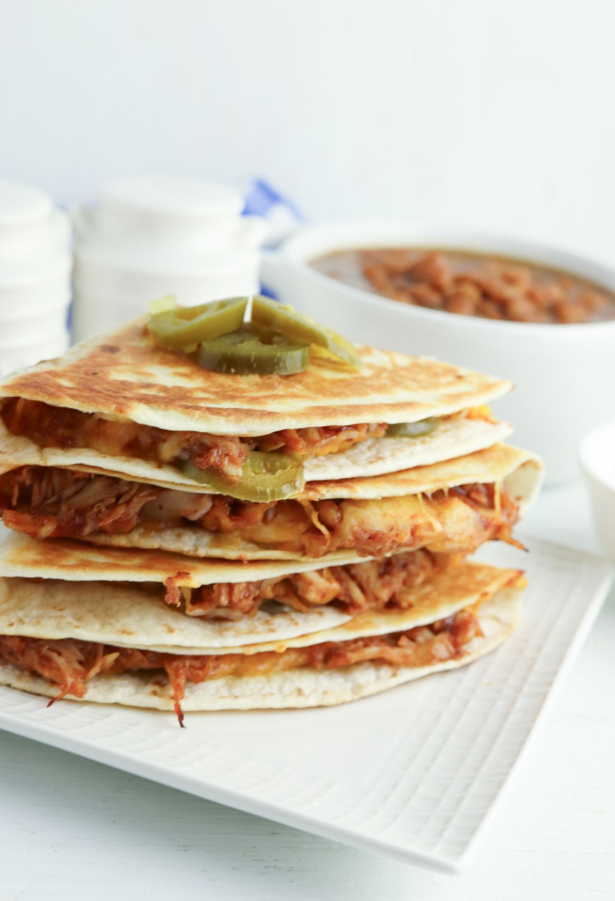 Stacked chicken quesadillas with pickled jalapeños on top, served on a rectangular white plate against a backdrop of more quesadillas and a side dish.