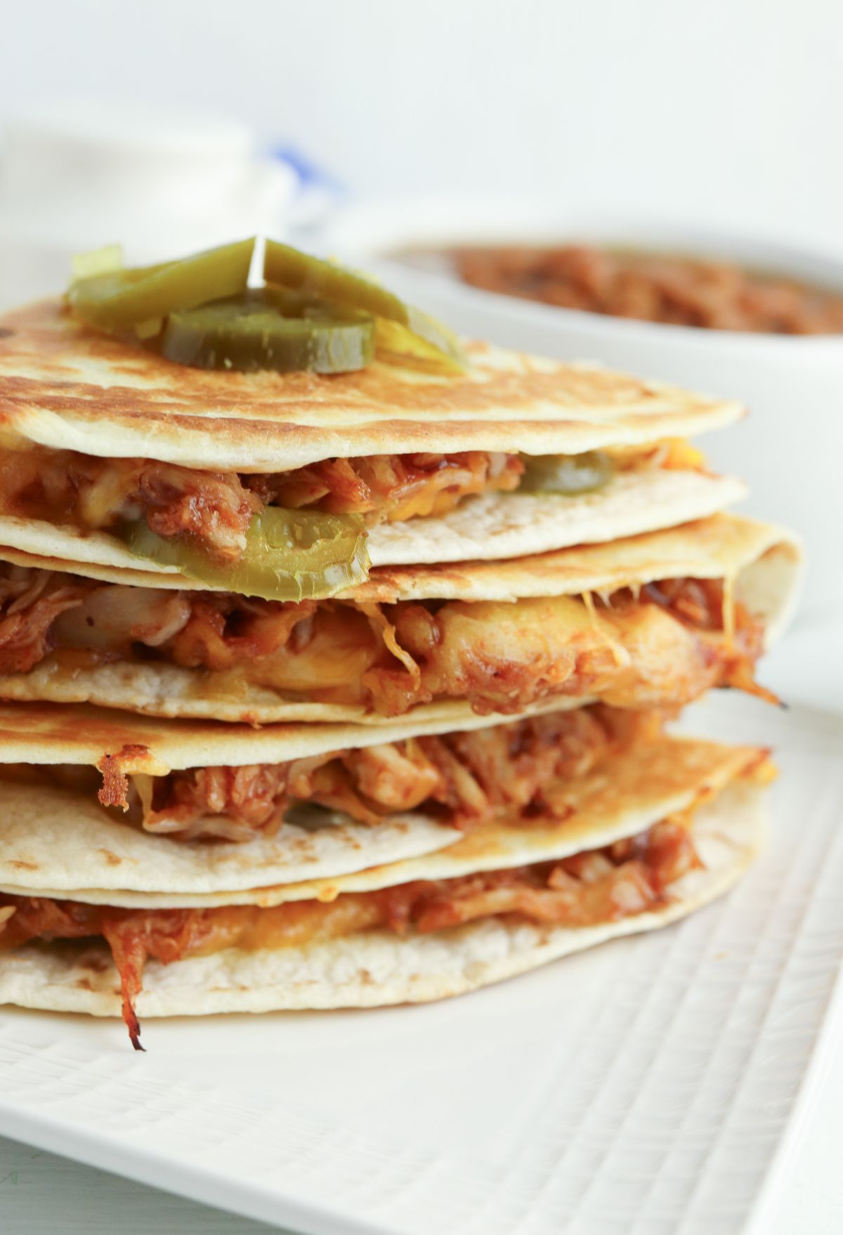 Stack of quesadillas filled with cheese and shredded chicken, topped with pickled jalapeños on a white plate.