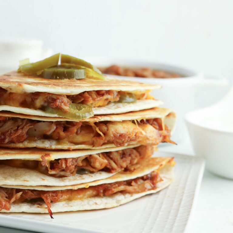 A stack of quesadillas filled with cheese and shredded meat, garnished with sliced pickles, on a white plate with a bowl of sauce in the background.