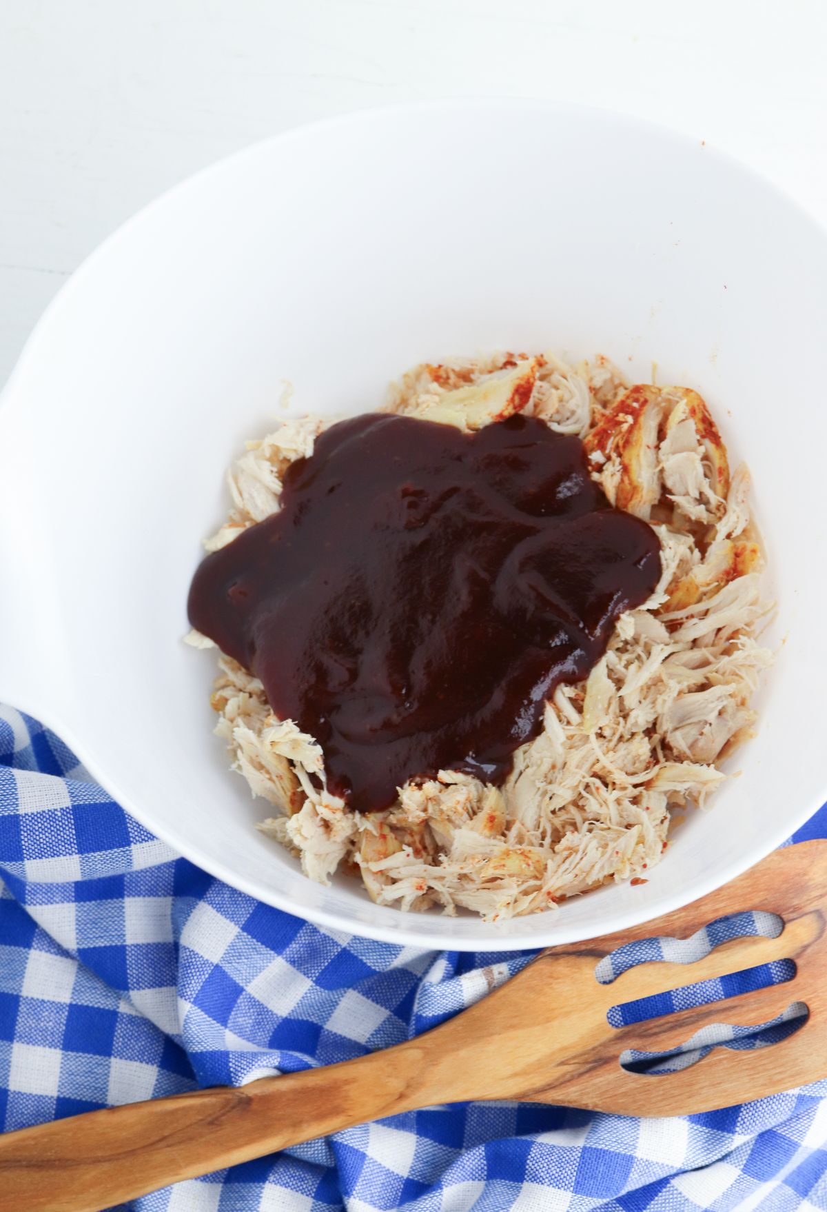 Shredded chicken topped with barbecue sauce in a white bowl, placed on a blue and white checkered cloth with a wooden fork to the side.